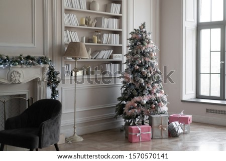 Christmas tree decorated with festive decor of luminous garlands, Assorted Baubles and decorative snow. Wrapped gift boxes under the Christmas tree. Room has a large library window and fireplace.
