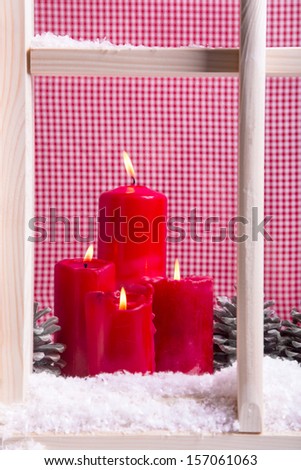 Indoor Christmas window sill decoration: red candles,snow and pine cones on chequered background.