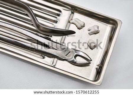 Chrome stainless steel medical tray with dental instruments (forceps and a cheek retractor) and three extracted molar teeth. Closeup horizontal photo.
