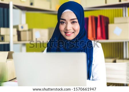 Muslim asian woman freelancer sme business online shopping working on laptop computer and checklist order with cardboard box on table at home - Business online shipping and delivery concept