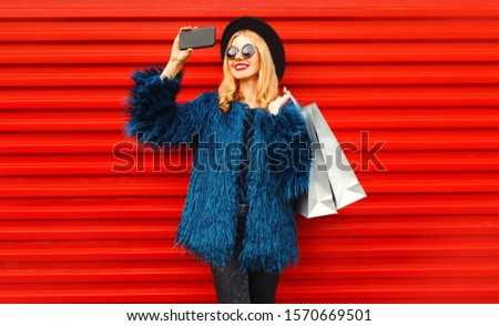 Attractive young smiling woman taking selfie picture by smartphone with shopping bags, stylish female model wearing blue faux fur coat, black round hat and sunglasses over red wall background