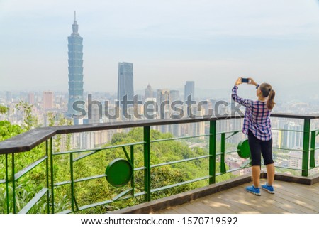 Young pretty female tourist taking pictures and enjoying view of Taipei from top of mountain. Awesome Taipei skyline, Taiwan. Wonderful cityscape. Skyscrapers and other modern buildings of downtown.