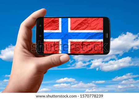 Norway flag on the phone screen. Smartphone in hand shows a flag on a background of the sky with clouds. Mobile photography.