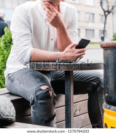 Photo of A young man in a white shirt and jeans sits at a table in a cafe on the street and looks at the phone