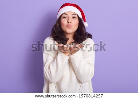 Picture of attractive young girl blowing kiss, putting hands together in front of herself, looking directly at camera, having tender facial expression, wearing hat for new year party. Rest concept.