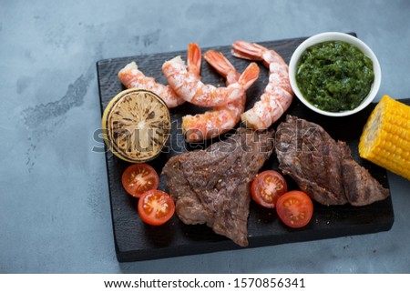 Surf and turf with marbled beefsteaks, shrimps, tomatoes, lemon and chimichurri on a black wooden serving board
