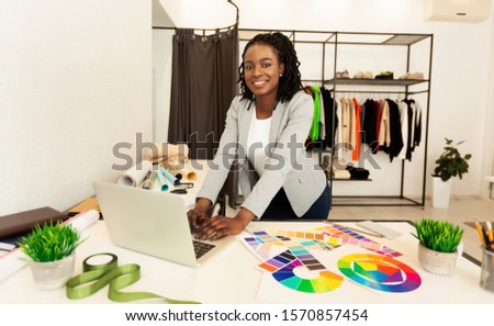 Cheerful Afro Tailor Girl Using Laptop Working On New Sewing Pattern Standing In Clothing Atelier. Fashion Entrepreneur Concept.
