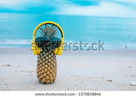 Ripe attractive pineapple in stylish mirrored sunglasses and gold headphones on sand against turquoise sea water. Tropical summer vacation concept.
