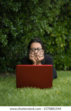 Brunette woman lying down on the grass of a park while watching the screen of her laptop. Natural Environment. Vertical Photography
