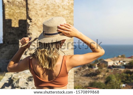 Caucasian tanned girl traveler in a hat and a bathing suit looks at the sea from afar.