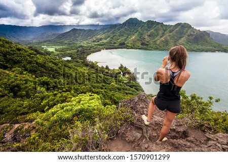 Rear view of a young woman on the top of a mountain, taking a picture with her smartphone of the beautiful landscape on the Caribbean coast near the sea 