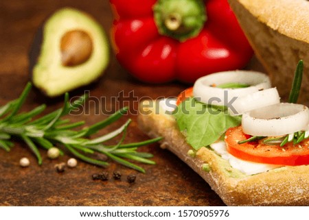 sandwich made of italian bread ciabatta on a brown table. onion, tomato, pepper, goat cheese, razmoin, basil and avocado.