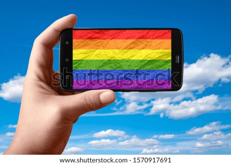 LGBT flag on the phone screen. Smartphone in hand shows a flag on a background of the sky with clouds. Mobile photography.