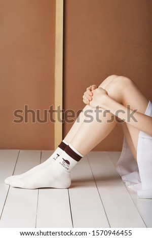 Cropped shot of a lady in a short skirt and white ribby socks with brown stripes, printed cartoon heroes and letterings "Awesome". The girl is sitting on the board floor against the brown wall. 