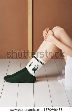 Cropped shot of a lady in a short skirt and white and green ribby socks with green stripes and printed cartoon heroes by the sides. The girl is sitting on the board floor against the brown wall. 
