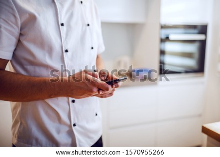 Cropped picture of caucasian chef in uniform standing in kitchen and using smart phone.