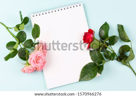 Sketchbook (Notepad) with a blank sheet of writing paper, pink and red roses close-up on a blue background. Women's business concept. happy birthday. Valentine's day. Space for text.