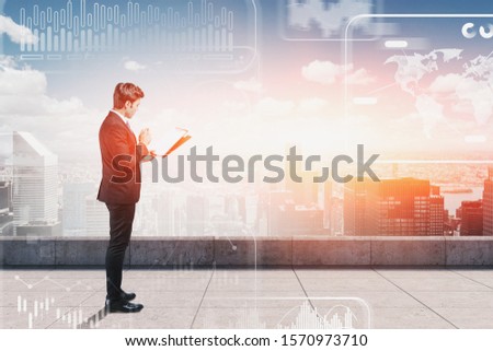 Young businessman with clipboard working in modern city standing on skyscraper roof with double exposure of blurry infographics interface. Concept of hi tech and stats. Toned image