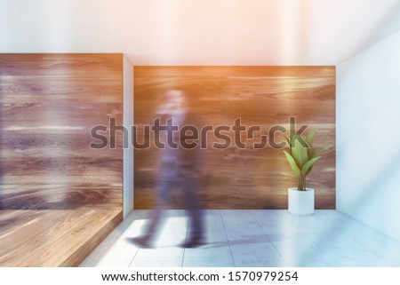 Blurry businessman walking in empty room with white and wooden walls, white tiled floor and big potted plant. Toned image