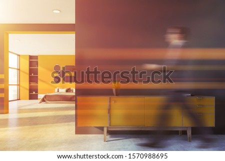 Blurry young man walking in modern bedroom with yellow and gray walls, concrete floor, gray king size bed with mirrors above it and yellow cabinet. Toned image