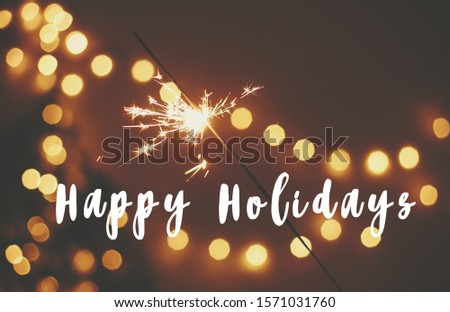 Happy Holidays text sign on glowing sparkler in hand on background of golden christmas tree lights in dark festive room. Fireworks burning. Seasons greeting card
