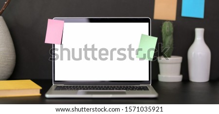 Cropped shot of trendy office room with mock-up laptop computer, office supplies and sticky note on black table with black wall background 