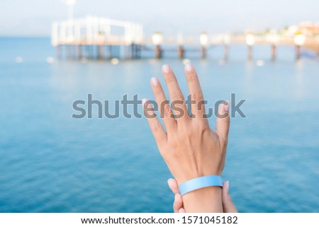Closeup view of beautiful white female hand wearing rubber wristband of hotel resort. Hand isolated st sunny summer bright blue sea water background. Horizontal color photography.