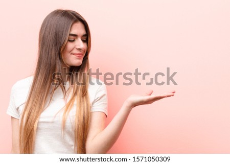 young pretty woman feeling happy and smiling casually, looking to an object or concept held on the hand on the side against pink wall