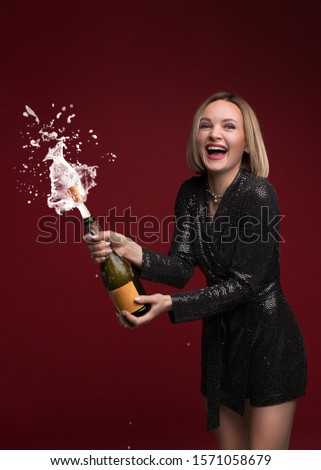 Beautiful young lady in black glitter sequins evening dress opening bottle of champagne, splashing champagne in her hands. Celebration Christmas, New Year's party.