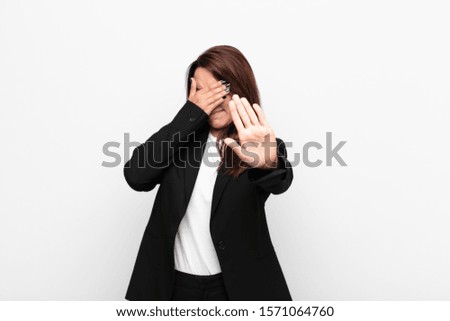 young pretty businesswoman covering face with hand and putting other hand up front to stop camera, refusing photos or pictures against white wall