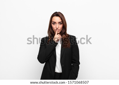 young pretty businesswoman looking serious and cross with finger pressed to lips demanding silence or quiet, keeping a secret against white wall