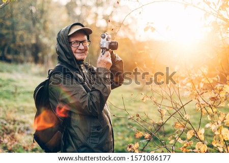 Handsome senior man photographer standing in autumn park and taking pictures with camera.