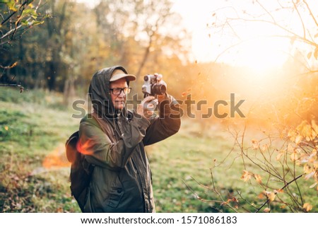 Old man photographer standing in autumn park and taking pictures with camera.