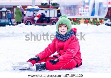 Happy model boy or girl is sitting on a city ice rink in skates and ski overalls against the background of Christmas decorations. Winter activities outdoors. Winter leisure for children
