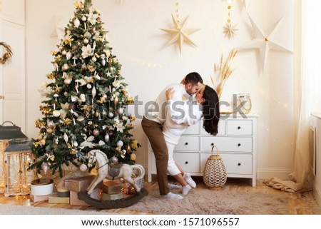 An attractive brunette girl in a white wool dress and a brutal man in a sweater hug and kiss on the background of a Christmas tree, a chest of drawers and decorative stars. happy family and Christmas.