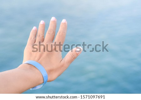 Closeup view of beautiful white female hand wearing blue rubber wristband. Hand isolated at blurry sea water bokeh background. Happy travel and all inclusive system of hotel resorts concept.