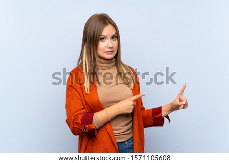 Teenager girl with coat over isolated blue background frightened and pointing to the side