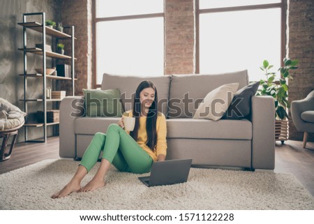 Full length photo of interested girl sit floor carpet use computer search internet movie hold cup with cacao wear yellow pullover green pants in house room