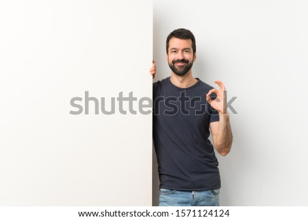 Young handsome man with beard holding a big empty placard showing ok sign with fingers