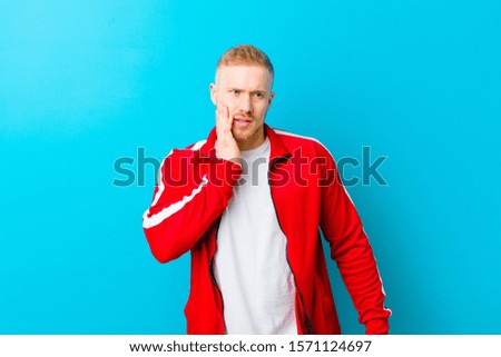 young blonde man wearing sports clothes holding cheek and suffering painful toothache, feeling ill, miserable and unhappy, looking for a dentist against blue background