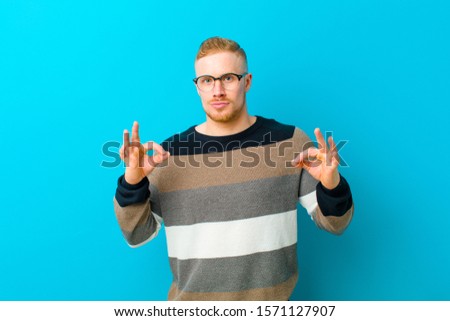 young blonde man feeling shocked, amazed and surprised, showing approval making okay sign with both hands against blue background