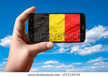 Belgium flag on the phone screen. Smartphone in hand shows a flag on a background of the sky with clouds. Mobile photography.