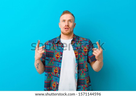 young blonde man wearing a print shirt looking shocked, amazed and open mouthed, pointing upwards with both hands to copy space