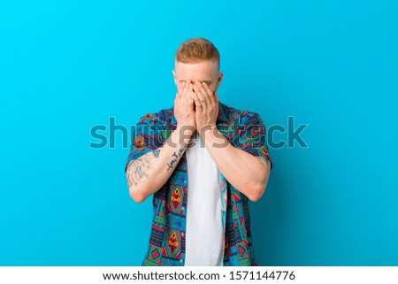 young blonde man wearing a print shirt feeling sad, frustrated, nervous and depressed, covering face with both hands, crying