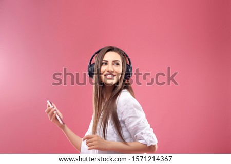 Beautiful young woman in wireless headphones listening to music using mobile phone and dancing on pink background. Girl uses wireless earphones