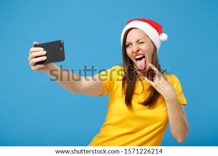 Crazy Santa girl in Christmas hat posing isolated on blue background. New Year 2020 celebration holiday concept. Mock up copy space. Doing selfie shot on mobile phone, depicting heavy metal rock sign