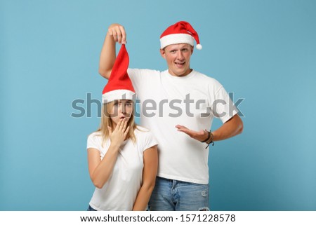 Confused young Santa couple friends guy girl in Christmas hat posing isolated on pastel blue background. Happy New Year 2020 celebration holiday concept. Mock up copy space. Covering mouth with hand