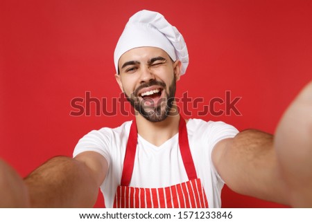 Close up of blinking young bearded chef cook baker man in striped apron toque chefs hat posing isolated on red background. Cooking food concept. Mock up copy space. Doing selfie shot on mobile phone