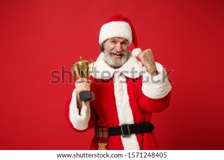 Elderly gray-haired mustache bearded Santa man in Christmas hat posing isolated on red background. New Year 2020 celebration holiday concept. Mock up copy space. Hold soccer cup, doing winner gesture