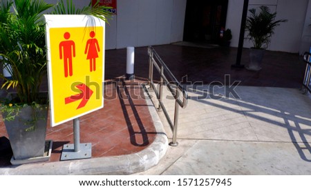Sunlight on surface of restroom sign on metal pole with stainless steel fence on the different level ground in parking area, Thai text in red label is mean danger zone